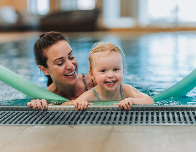 Mutter und Tochter spielen lachend im Pool mit einer Schwimmnudel.