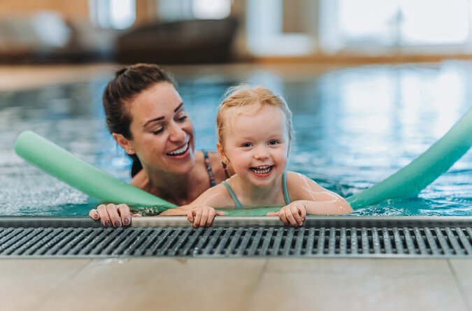 Mutter und Tochter spielen lachend im Pool mit einer Schwimmnudel.