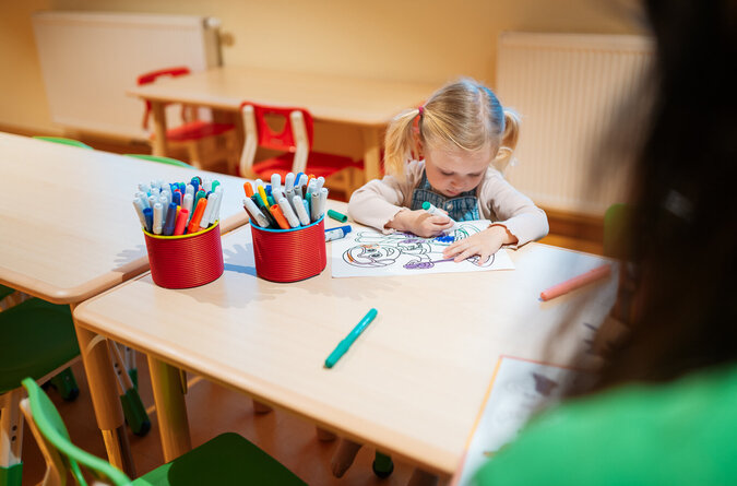 Ein Mädchen malt mit bunten Stiften auf Papier an einem Tisch.