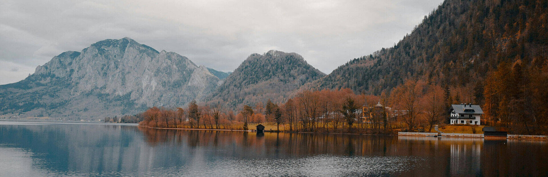 Herbstliche Landschaft mit ruhigem Bergsee, bewaldeten Hängen und einem Haus am Ufer, umgeben von den Alpen.