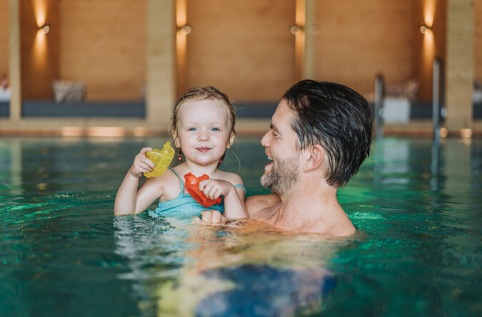 Vater und Tochter lachen gemeinsam beim Baden im Pool eines Familienhotels.