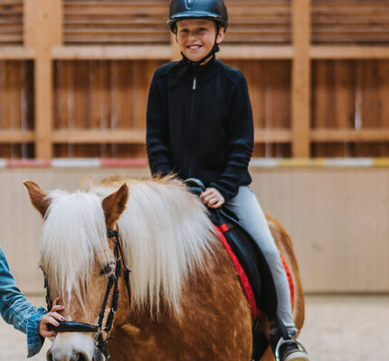 Ein Mädchen reitet auf einem Pferd, das von einer Reitlehrerin mit einem Seil geführt wird.